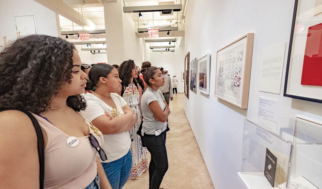 Group of people gathered in El Museo looking at a wall filled with framed pictures.