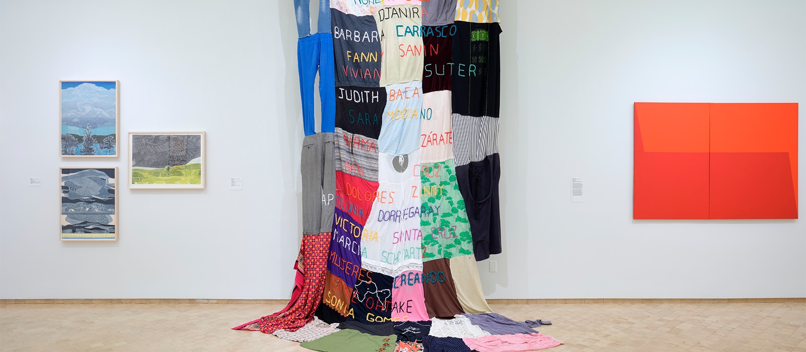 Carolina Caycedo. Muxeres en mi, 2019. Clothing and embroidery, 144 x 72 in. Gift of the Acquisitions Committee, 2022. Installation view at Something Beautiful: Reframing La Colección, El Museo del Barrio, 2023. Also displayed works by Myrna Báez (left) and Carmen Herrera (right). Photo On White Wall.