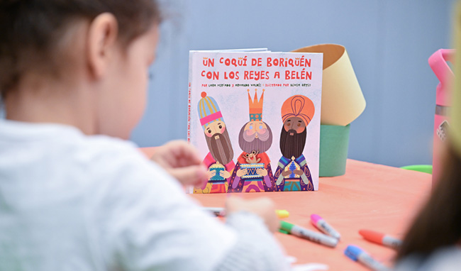 A child sits at a table, partially obscuring a book about the Three Kings, while engaging in a coloring activity.