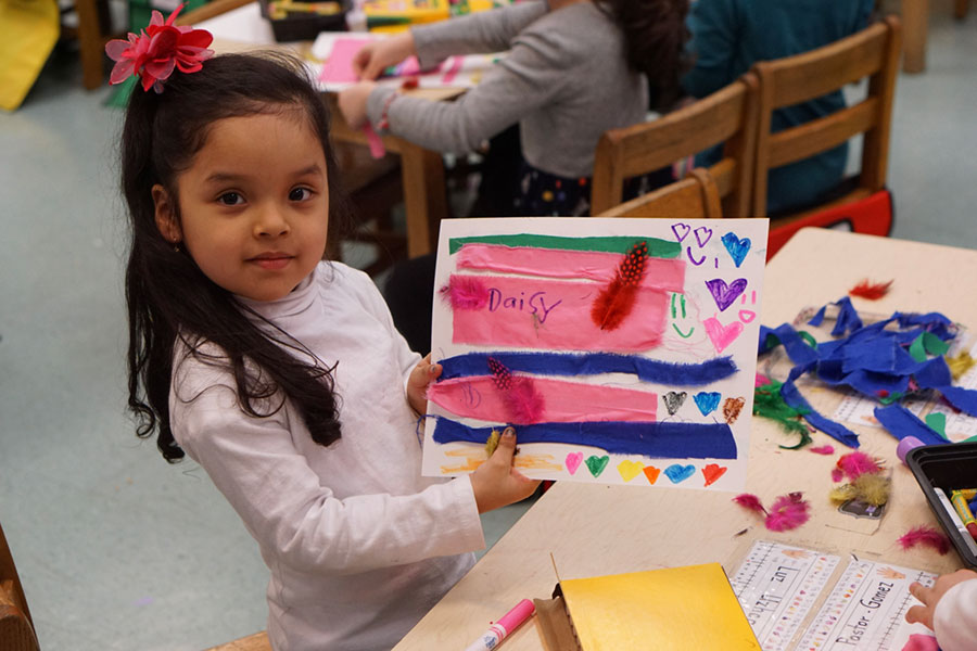 A young girl proudly displays her colorful mixed-media artwork, which features feathers, fabric scraps, and hand-drawn hearts.